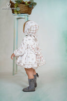 Baby wearing a white dress with deer print, matching bonnet, and ruffle details from DBB Collection AW25.