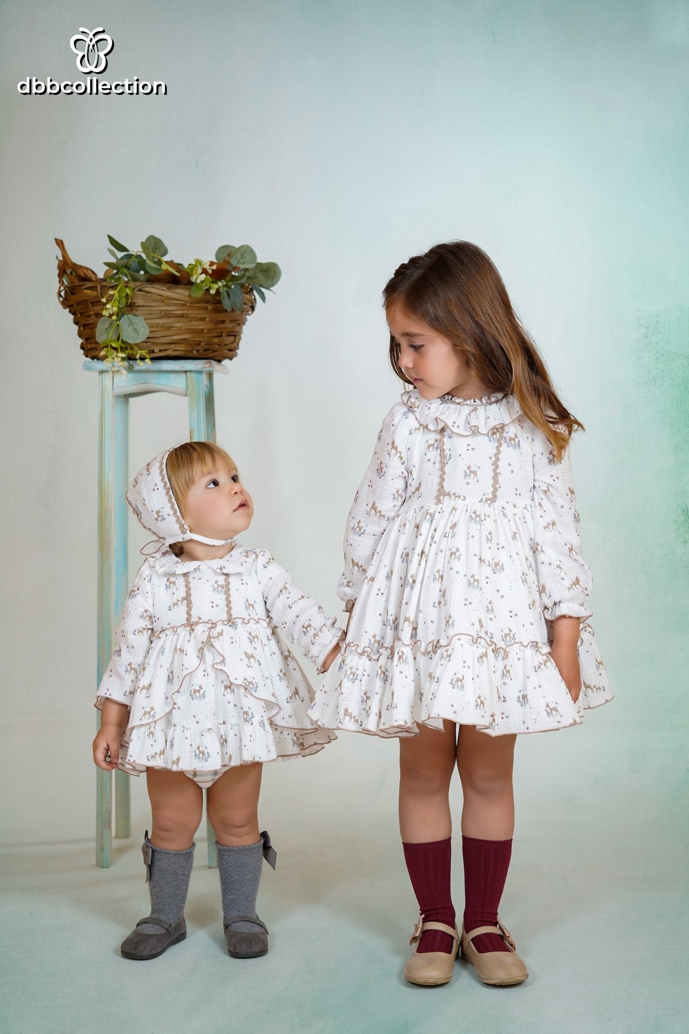 Baby wearing a white dress with deer print, matching bonnet, and ruffle details from DBB Collection AW25.