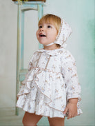 Baby wearing a white dress with deer print, matching bonnet, and ruffle details from DBB Collection AW25.