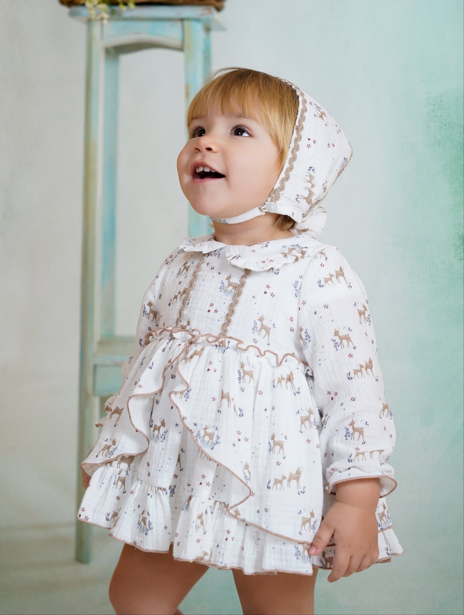 Baby wearing a white dress with deer print, matching bonnet, and ruffle details from DBB Collection AW25.