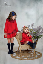 Girl wearing red long-sleeved holiday dress with navy gingham ruffle collar and bow, from DBB Collection AW25.