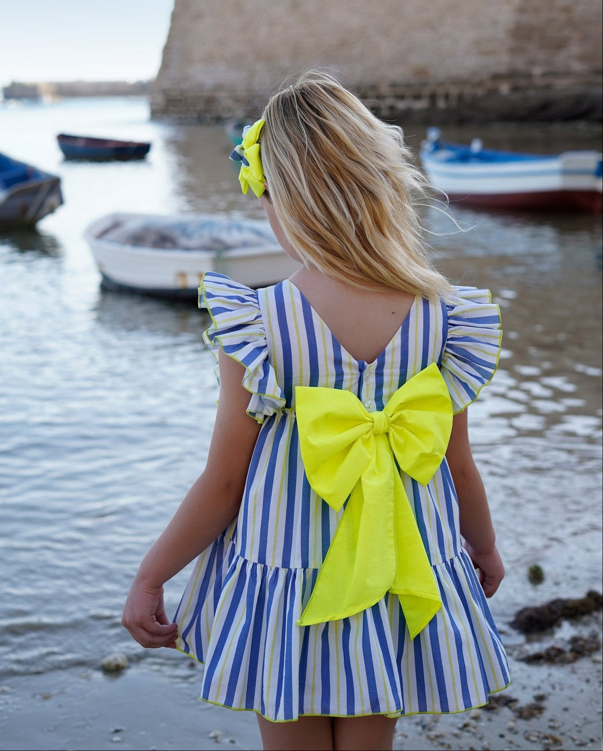 A young girl with blonde hair is seen from behind, wearing a blue and white striped dress with a large neon yellow bow on the back and a matching yellow hair clip, standing on a beach with water and a stone fort in the background.