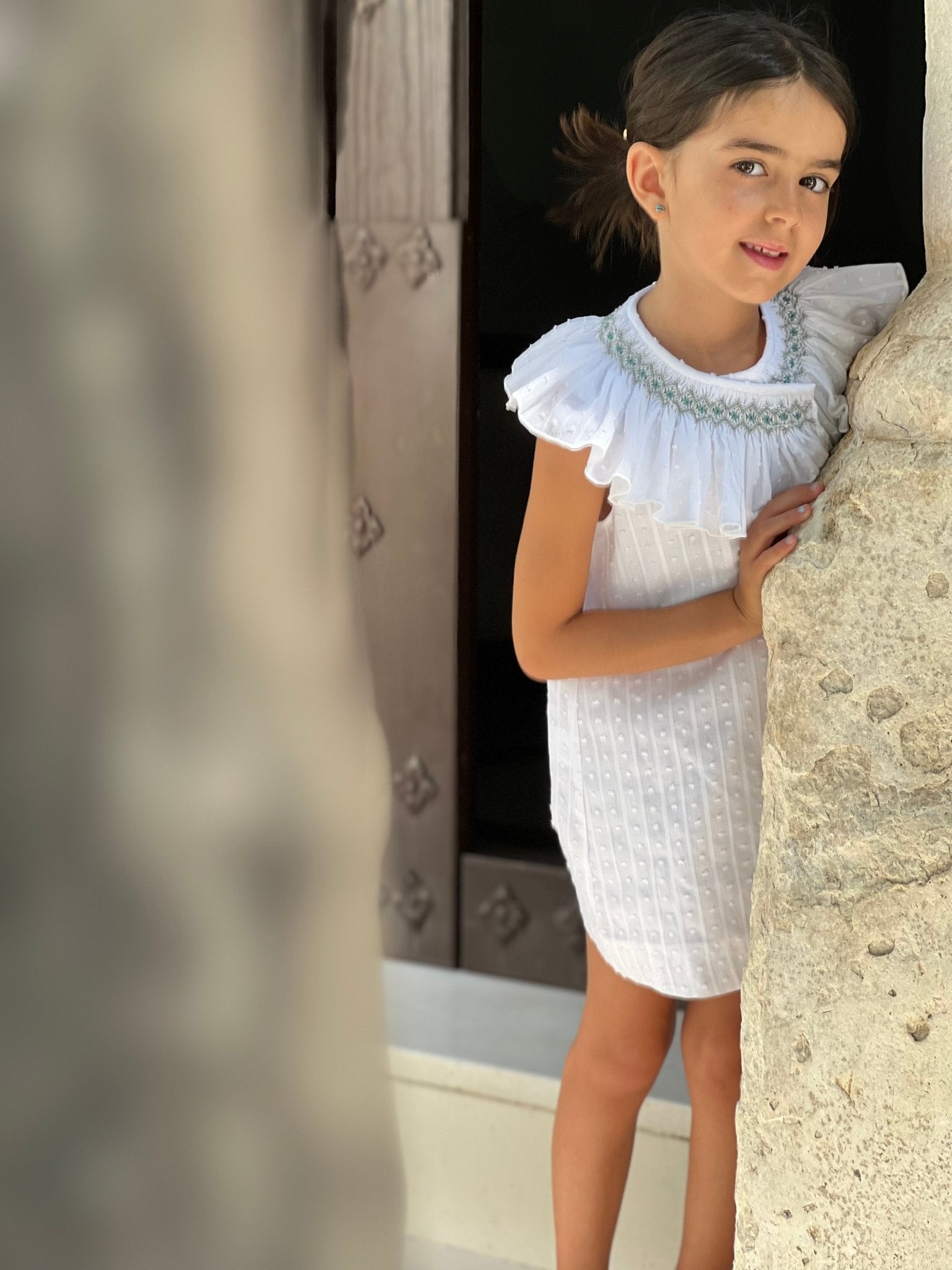 Young girl in a white smocked collar spanish dress standing against a stone wall by El Copo for SS26