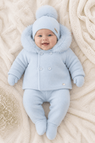 A baby lying on a cream knit blanket, wearing a light blue winter outfit with a hooded jacket, matching hat with pom-pom, and mittens.