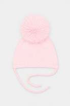 A light pink knit baby beanie hat with a fluffy pom-pom on top and matching tie strings, shown against a plain light gray background.