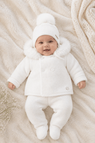 A baby lying on a soft cream blanket wearing a white knit winter outfit with a fluffy faux-fur trimmed hood, matching pom-pom beanie, and booties.