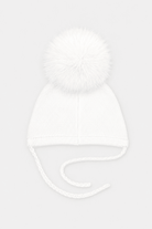 A white knit baby beanie hat featuring a large fluffy pom-pom on top and matching white ear tie strings, shown against a light gray background.