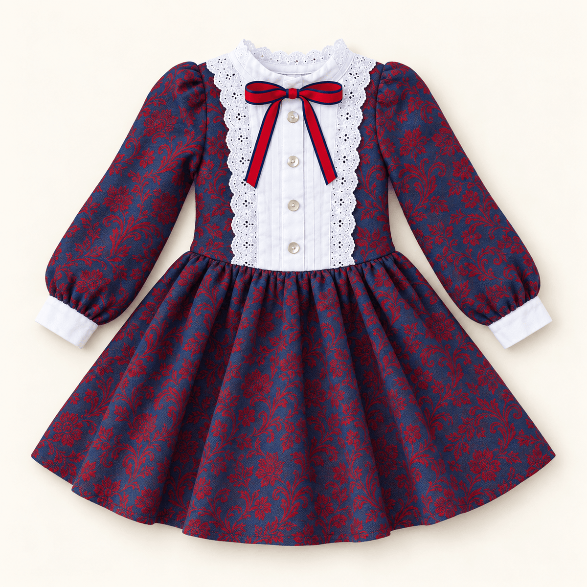 A long-sleeve girls dress with a navy and red damask pattern, featuring a white lace-trimmed yoke, white button-front placket, and a red bow at the collar, shown on a light cream background.