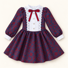 A long-sleeve girls dress with a navy and red damask pattern, featuring a white lace-trimmed yoke, white button-front placket, and a red bow at the collar, shown on a light cream background.