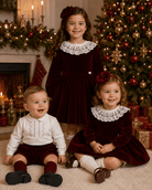 Three children dressed in matching Christmas outfits, featuring deep burgundy velvet dresses and tops with white lace collars, posed in front of a decorated Christmas tree and fireplace.