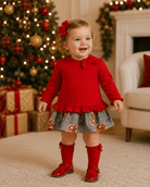 A toddler girl wearing a festive red long-sleeve top with a ruffled hem, a patterned gingham skirt with autumn motifs, red knee-high socks, and red shoes, standing in a cozy living room decorated for Christmas.