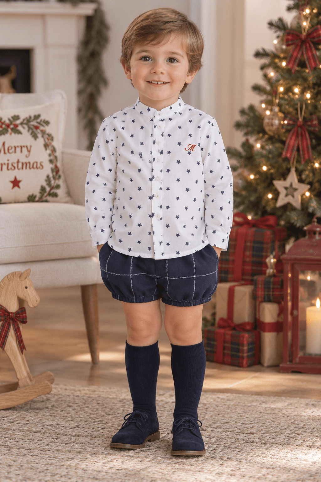 A young boy stands in a cozy holiday living room wearing a white long-sleeve button-up shirt with small navy polka dots, navy plaid shorts, navy knee-high socks, and navy shoes. A Christmas tree, wrapped gifts, and a fireplace are visible in the background.