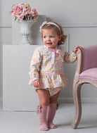 Baby girl in Rochy AW25 Rococo multi colour dress & knickers set standing in a room with a vase of flowers and a chair.