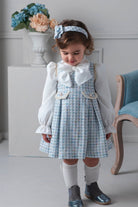 Young girl in a checkered dress with a white blouse and headband, standing indoors.