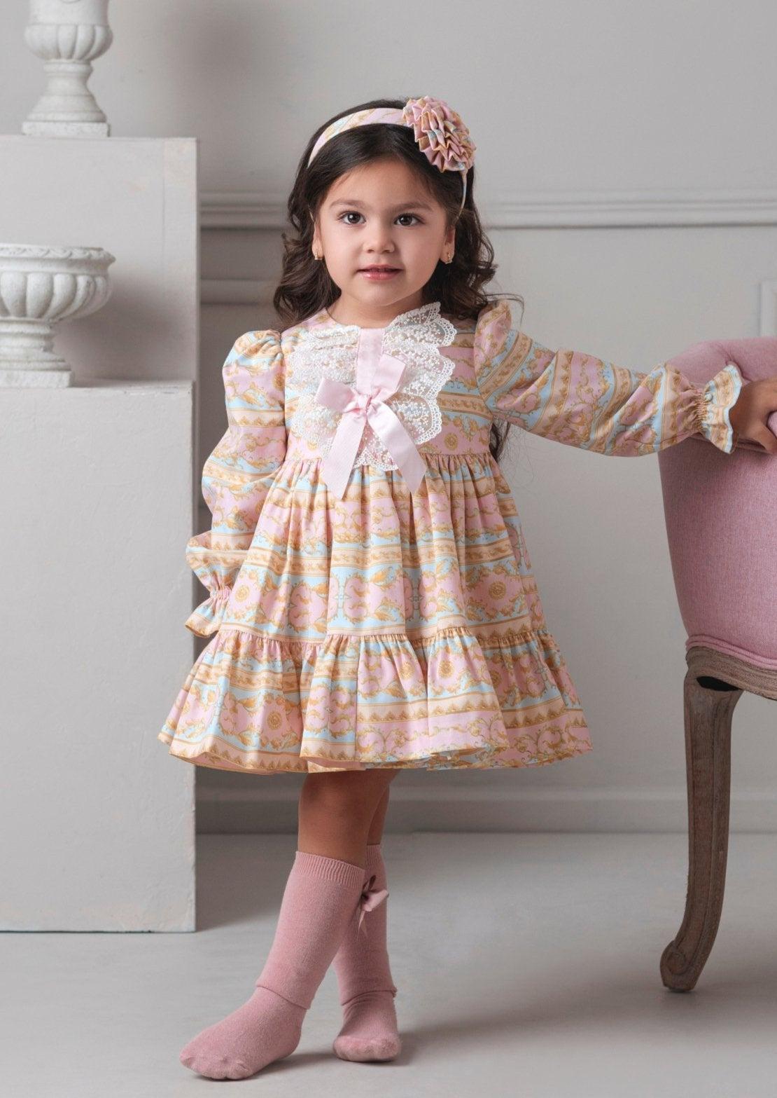 Young girl in a patterned Rochy AW25 dress standing in a room with decorative elements.