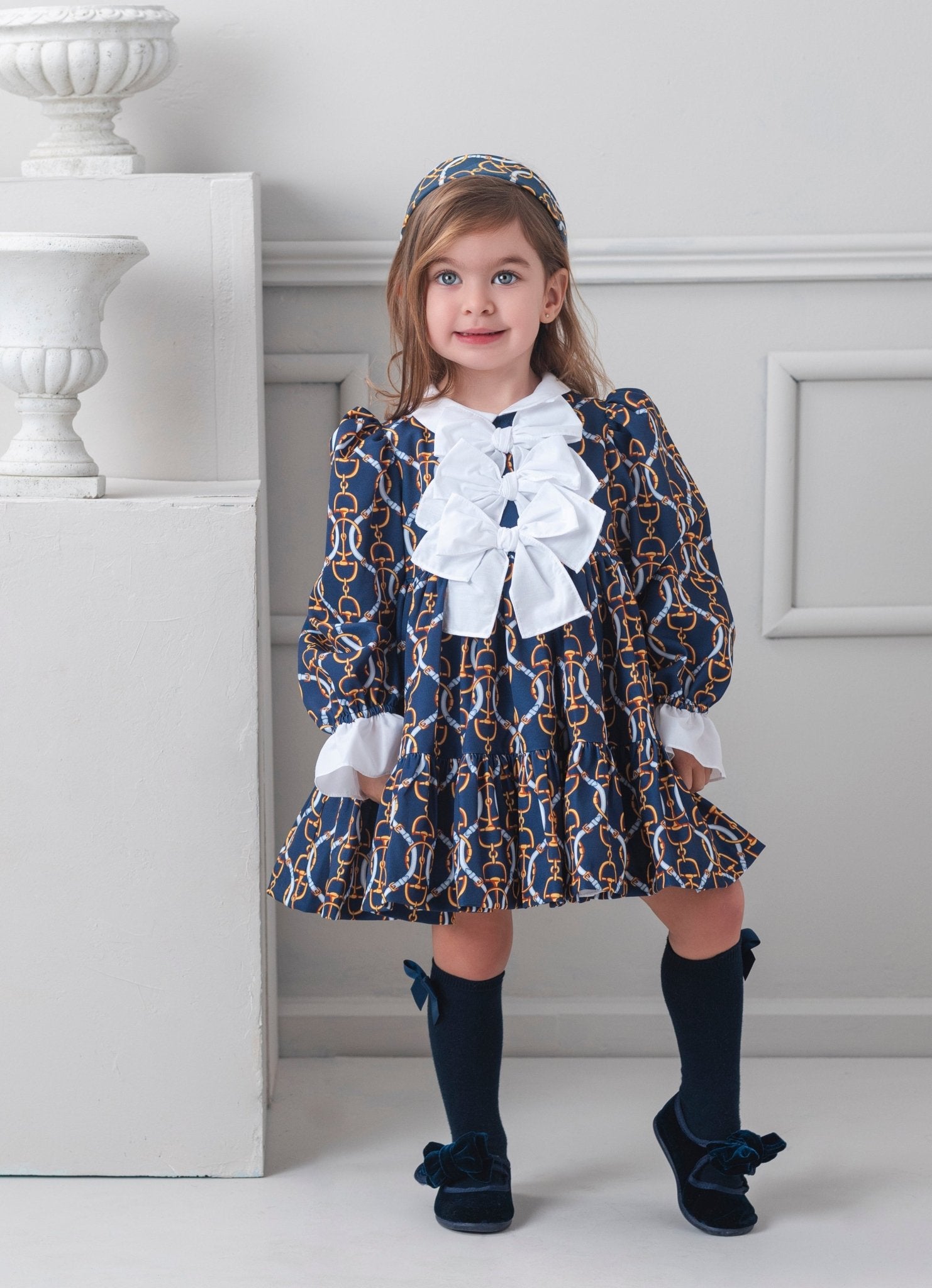 Young girl in a patterned Rochy AW25 Vintage Chains Navy and Gold dress with white bows standing in a room with decorative elements.