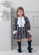 Young girl in a patterned Rochy AW25 Vintage Chains Navy and Gold dress with white bows standing in a room with decorative elements.
