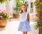 A young girl stands in a sunlit outdoor courtyard wearing a sleeveless blue and pink plaid dress with a white ruffled collar and bow detail. Pink flowers and greenery frame the scene.