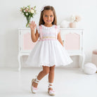 Young girl in a Sonata white smocked dress with pink accents waving in a bright room.