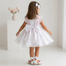 Young girl wearing a white Sonata dress with pink accents and a large bow, standing against a plain background.