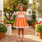 Young girl in an smocked Sonata orange dress standing in a garden with orange trees and potted plants.
