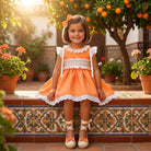 Young girl in an Sonata smocked orange dress with white lace trim sitting on a decorative ledge.